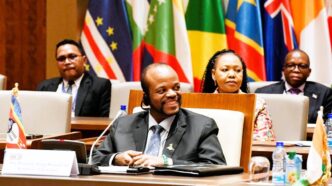 His Majesty King Mswati III smiles during the OACPS resource mobilisation session as Minister of Foreign Affairs Pholile Shakantu looks on from behind.