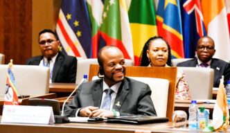 His Majesty King Mswati III smiles during the OACPS resource mobilisation session as Minister of Foreign Affairs Pholile Shakantu looks on from behind.