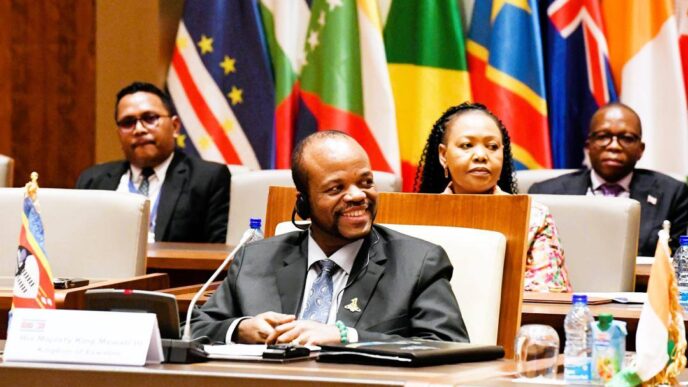 His Majesty King Mswati III smiles during the OACPS resource mobilisation session as Minister of Foreign Affairs Pholile Shakantu looks on from behind.